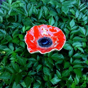 May include: A vibrant, handcrafted ceramic poppy flower sculpture with a glossy red petal design and a dark blue center. The flower is set against a backdrop of lush, green foliage, creating a striking garden accent.