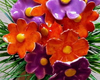 Könnte beinhalten: Ein Strauß aus Keramikblumen in den Farben Lila, Orange und Gelb. Die Blumen haben eine glänzende Oberfläche und sind in einem Bett aus grünem Gras angeordnet.
