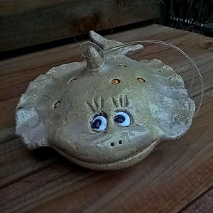 Könnte beinhalten: Eine Keramikfigur eines grauen und weißen Fisches mit großen blauen Augen und orangefarbenen Flecken. Der Fisch hat einen lächelnden Mund und hängt an einer Schnur.