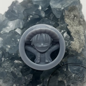 Può includere: Una statuetta grigia stampata in 3D di un personaggio dei cartoni animati con un cappello di paglia e un grande sorriso. La statuetta è seduta su un letto di cristalli blu.