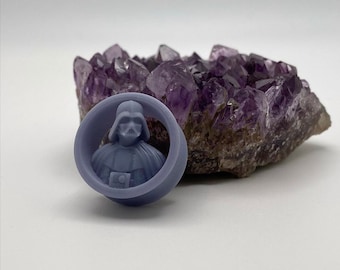 May include: A dark gray 3D printed Darth Vader bust inside a round, hollow, gray frame. The frame is sitting on a cluster of purple amethyst crystals.