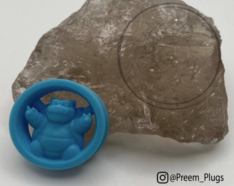 May include: A blue and white ear gauge featuring a 3D Snorlax Pokemon character. The gauge is on a light brown rock.
