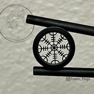 Könnte beinhalten: Schwarzer runder Ohrstecker mit einem ausgeschnittenen Design eines schwarzen achtzackigen Sterns. Der Ohrstecker liegt auf einem schwarzen zylindrischen Objekt.