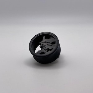May include: A black, circular plug with a detailed relief of a stylized face. The plug has a rounded base and a flared, cylindrical top. The face is a prominent feature, with sharp angles and defined features.