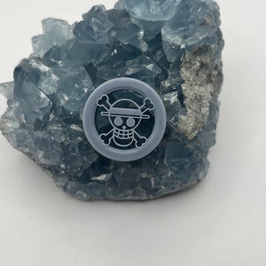 Puede incluir: Un amuleto de plástico gris con un diseño de calavera y huesos cruzados blancos. La calavera lleva un sombrero de paja.