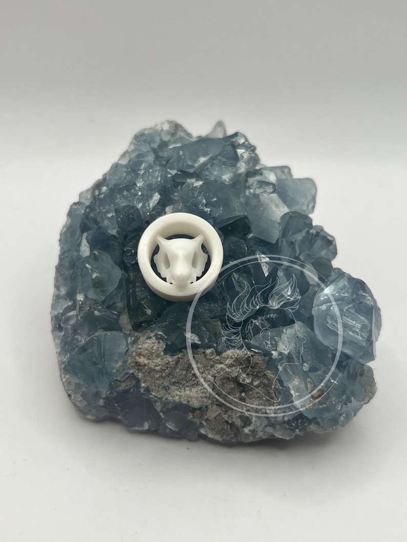 May include: White ceramic plug with a detailed animal face design, resting on a bed of blue crystals.