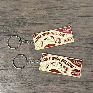 May include: Two rectangular keychains featuring a cream-coloured background with maroon text and graphics. The text reads ONE WISH WILLOW, AMAZE YOUR FRIENDS!, and You only get ONE WISH!. Each keychain includes a metal ring and a short chain attachment.