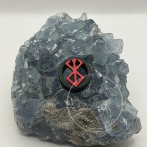 Può includere: Un anello nero con un simbolo rosso, appoggiato su una formazione di cristallo blu-grigio. Il simbolo è un disegno stilizzato a forma di croce. L'anello è il punto focale, sullo sfondo strutturato del cristallo.
