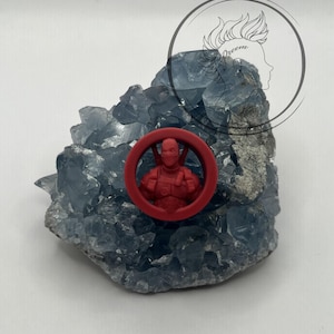 May include: Red plastic keychain featuring a cartoon character in a red and black suit. The keychain is on a blue crystal rock.