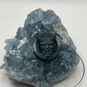 Pu&ograve; includere: Misura per orecchio circolare nera con un design dettagliato di un viso demoniaco. La misura &egrave; mostrata su una roccia di cristallo blu.