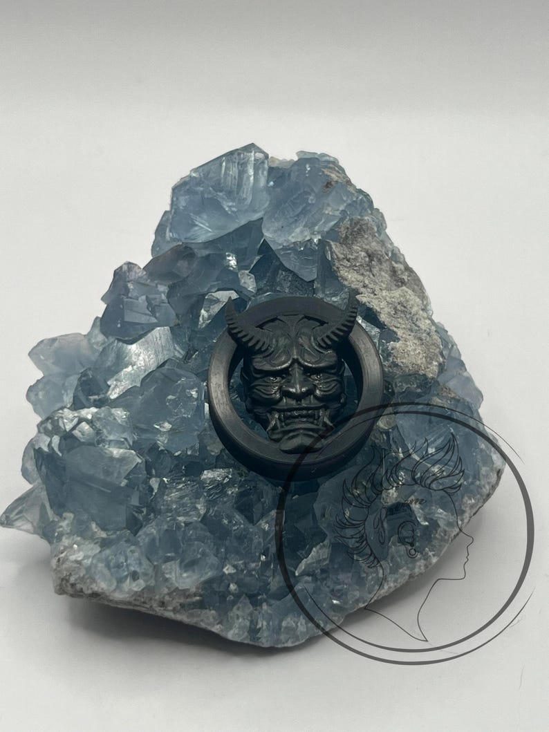 Pu&ograve; includere: Misura per orecchio circolare nera con un design dettagliato di un viso demoniaco. La misura &egrave; mostrata su una roccia di cristallo blu.