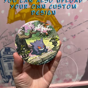 May include: A round button featuring a purple Gengar character in a Japanese garden scene with cherry blossoms. The button has the text "YOU CAN ALSO UPLOAD YOUR OWN CUSTOM DESIGN" in blue.