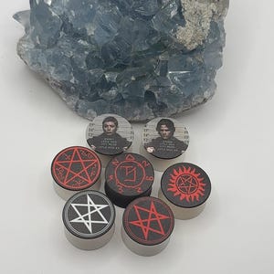 May include: A collection of six circular ear plugs featuring various graphic designs, including two mugshot portraits with text reading Little Rock City Police, a red pentagram, a red sun-like symbol, and two different white and red star emblems.
