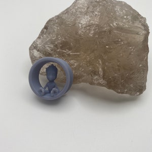 May include: A light purple ear gauge with a small, detailed figurine of a character sitting inside the gauge.