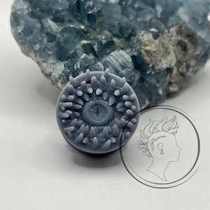 May include: A gray, circular, eye-shaped plug with a textured, spiky outer edge. The plug is made of a smooth, matte material.