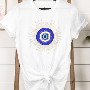 May include: White t-shirt with a blue and white evil eye design with gold rays radiating from the eye.