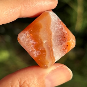 May include: A square-shaped, polished gemstone with a mix of orange, white, and red hues. The stone is held between fingers, showcasing its natural patterns and textures. The gemstone is a natural crystal.