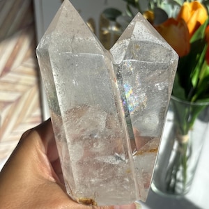 May include: Two clear quartz crystal points are held in a hand. The crystals are transparent with visible inclusions and a rainbow sheen. The crystals are a light, neutral color and are displayed in natural light.