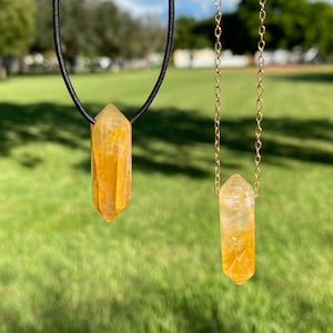 May include: Two necklaces with citrine crystal pendants. One pendant hangs from a black cord, the other from a gold chain. The crystals are a translucent yellow-orange color, with visible internal patterns. The background is a green field under a blue sky.
