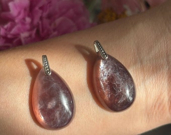 May include: Two teardrop-shaped pendants with a silver bail. The pendants have a translucent, reddish-brown hue with internal white veining. The background features pink flowers.