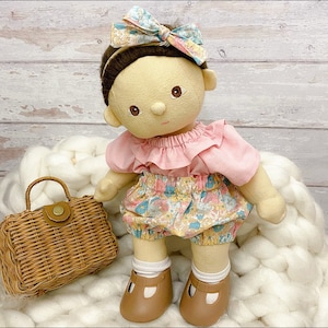Pode incluir: Uma boneca feita à mão com uma blusa rosa claro com babados, shorts de estampa floral e uma bolsa de vime marrom. A boneca está usando uma faixa de cabeça floral e sapatos marrons.