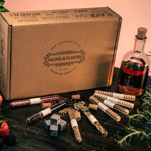 Puede incluir: Una caja de cartón marrón con el texto "FLAVOR OAK INDIVIDUAL PREMIUM BOTANICALS" impreso en la parte superior. La caja está abierta y contiene una variedad de pequeños tubos de vidrio llenos de diferentes especias y hierbas. También hay algunas piedras cuadradas de color gris oscuro y una botella de vidrio transparente con un tapón de corcho llena de un líquido oscuro. Los artículos están dispuestos sobre una superficie de madera.