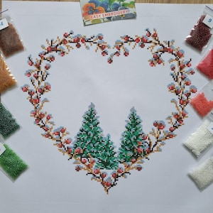 May include: A white fabric canvas featuring a cross-stitch pattern of a heart-shaped wreath made of brown branches, red berries, and two green pine trees at the base. Small plastic bags containing colored beads are arranged around the design.