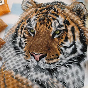 May include: Close-up of a tiger's face, created with diamond painting beads. The tiger's fur is a mix of orange, brown, and black, with white stripes. The tiger's eyes are green, and its nose is pink. The background is white.