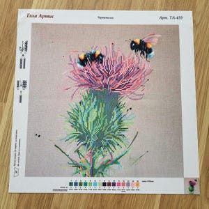 May include: A cross-stitch pattern on fabric featuring a thistle flower in shades of green and pink with two bumblebees. The top includes the text "Тэла Артис", "Чертополох", and "Арт. ТА-459". The bottom displays a color key with fifteen numbered circles.