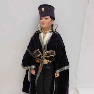 May include: A doll wearing a purple velvet robe with gold trim and a black hat with a gold emblem. The doll has a white shirt and black pants. The doll is wearing black boots.