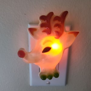 May include: A white plastic reindeer nightlight with red antlers and green hooves. The light is plugged into an outlet and is lit up.
