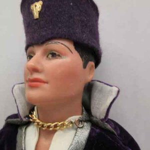 May include: A purple velvet hat with a gold emblem, worn by a doll with a white shirt and a gold chain necklace. The doll is wearing a purple velvet jacket with a silver trim.