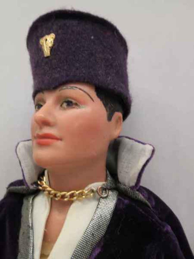 May include: A purple velvet hat with a gold emblem, worn by a doll with a white shirt and a gold chain necklace. The doll is wearing a purple velvet jacket with a silver trim.