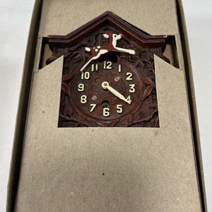 May include: A miniature brown cuckoo clock with a deer figurine on top, displayed in its original cardboard box. The clock face features Roman numerals and decorative floral carvings. The clock hands are white and the deer is white with brown spots.