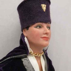 May include: A purple velvet doll with a purple felt hat with a gold emblem. The doll is wearing a white shirt and a gold chain necklace.
