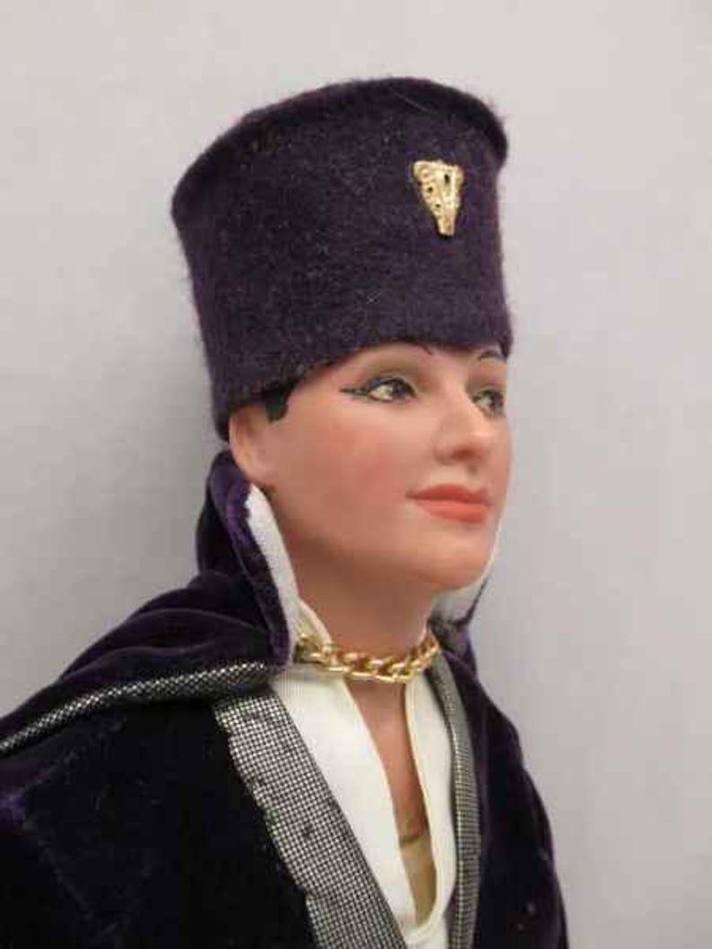 May include: A purple velvet doll with a purple felt hat with a gold emblem. The doll is wearing a white shirt and a gold chain necklace.