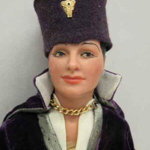 May include: A doll wearing a purple velvet jacket with a white shirt underneath. The doll is wearing a purple hat with a gold ornament on top. The doll has a gold chain necklace.