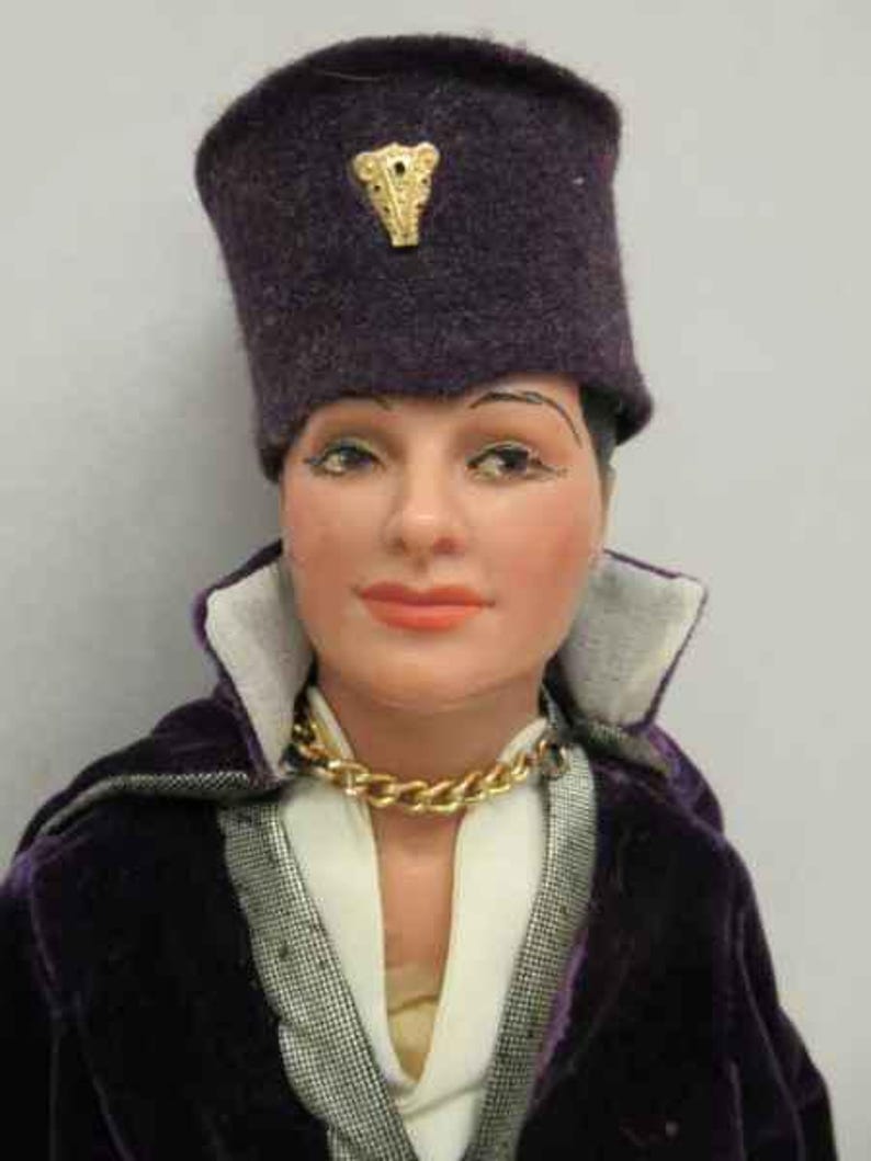 May include: A doll wearing a purple velvet jacket with a white shirt underneath. The doll is wearing a purple hat with a gold ornament on top. The doll has a gold chain necklace.