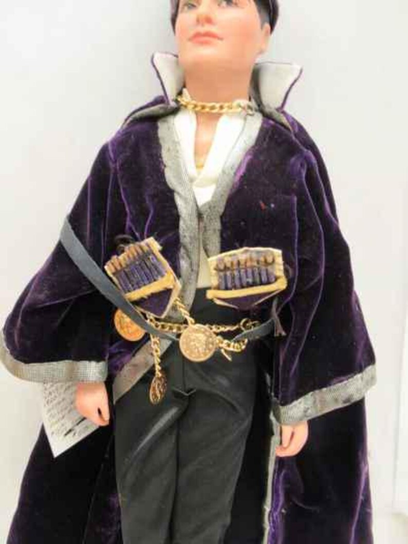 May include: A purple velvet robe with gold trim and a gold chain necklace. The robe has a decorative design on the front and is worn over a white shirt and black pants.