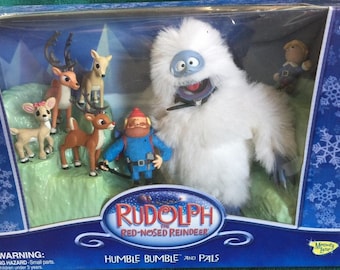 May include: A boxed set of Rudolph the Red-Nosed Reindeer figures, including Bumble, reindeer, and other characters. The box is blue with snowflake designs and the text "Rudolph the Red-Nosed Reindeer Humble Bumble and Pals."