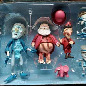 May include: A set of animated character figurines in a plastic display case. The figures include a Santa Claus-like character, a snowman-like figure, and other whimsical characters. The color palette includes red, blue, white, and pink.