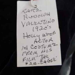 May include: A white tag with black handwritten text that reads "KARE RODOLPH VALENTINO 1920's Holly wood ACTOR in COSTUMZ FROM HIS FILM "THE 250-EAGLE"