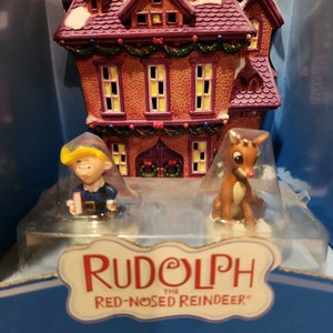 May include: A Rudolph the Red-Nosed Reindeer collectible set featuring a miniature porcelain house with purple roof, a small elf figurine, and a reindeer figurine with a red nose, all housed in original packaging with the text Rudolph the Red-Nosed Reindeer.