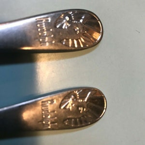 K&ouml;nnte beinhalten: Zwei Edelstahl-L&ouml;ffel mit einem dekorativen Design am Ende. Das Design beinhaltet das Wort "Regina" und einen stilisierten Sonnenaufgang mit einem Vogel oder Insekt in der Mitte. Die L&ouml;ffel liegen auf einer hellblauen Oberfl&auml;che.