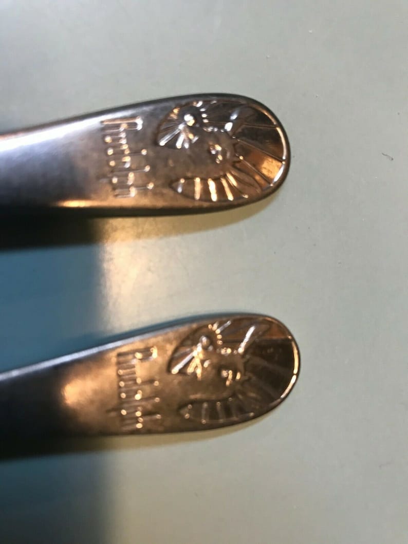 K&ouml;nnte beinhalten: Zwei Edelstahl-L&ouml;ffel mit einem dekorativen Design am Ende. Das Design beinhaltet das Wort "Regina" und einen stilisierten Sonnenaufgang mit einem Vogel oder Insekt in der Mitte. Die L&ouml;ffel liegen auf einer hellblauen Oberfl&auml;che.