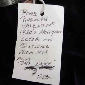 May include: A white paper tag with black handwritten text that reads "RARE RUDOLPH VALENTINO 1920's Hollywood ACTOR IN Costume FROM HIS FILM "THE EAGLE" 258".