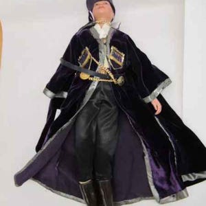 May include: A porcelain doll wearing a purple velvet coat with gold trim and a black hat. The doll is wearing black pants and black boots.
