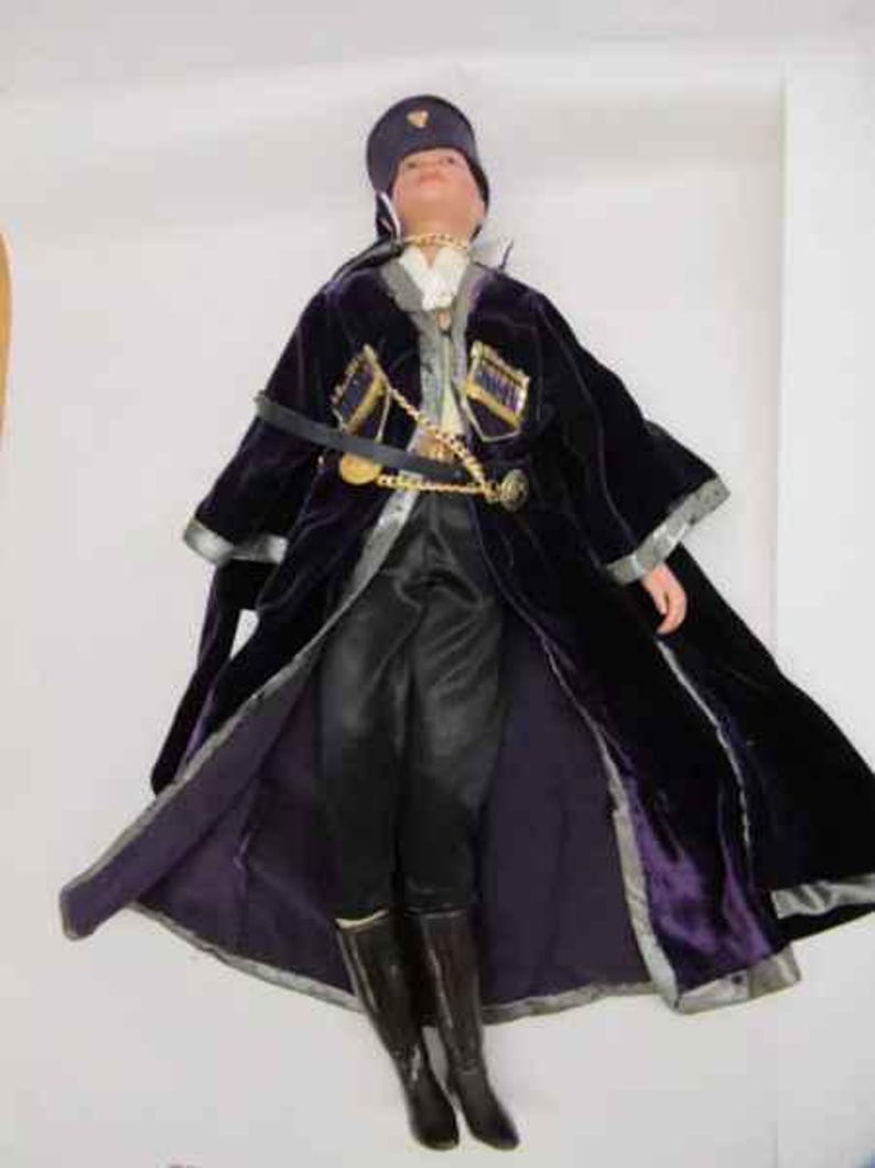 May include: A porcelain doll wearing a purple velvet coat with gold trim and a black hat. The doll is wearing black pants and black boots.