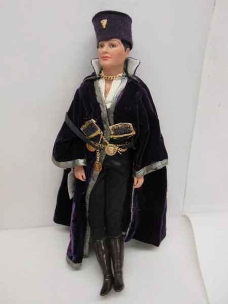 May include: A doll wearing a purple velvet robe with gold trim and a black hat with a gold emblem. The doll has a white shirt and black pants. The doll is wearing black boots.