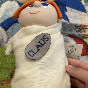 May include: A plush doll with a round head, orange hair, and blue eyes, wrapped in a white knitted blanket. A gray oval tag with the name "CLAUS" is attached to the blanket. A white tag with a logo is visible.